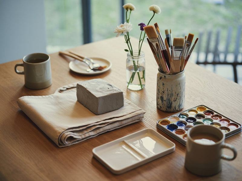 A studio worktable set before a class: a folded apron, a block of clay, a jar of paintbrushes, a watercolour palette, flower stems in a jar of water and two mugs of coffee.